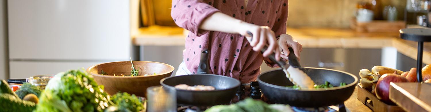Une personne cuisine devant une plaque de cuisson, mélange des légumes dans une poêle, entourée sur le plan de travail de lég