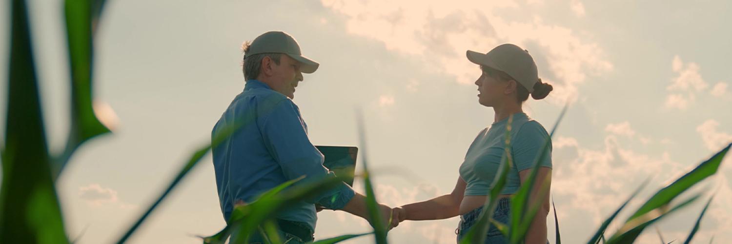 Deux personnes se serrent la main au milieu d’un champ de cultures, avec de la végétation au premier plan et un ciel lumineux