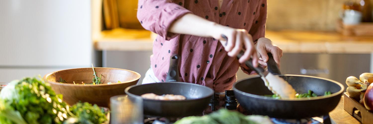 Une personne cuisine devant une plaque de cuisson, mélange des légumes dans une poêle, entourée sur le plan de travail de lég