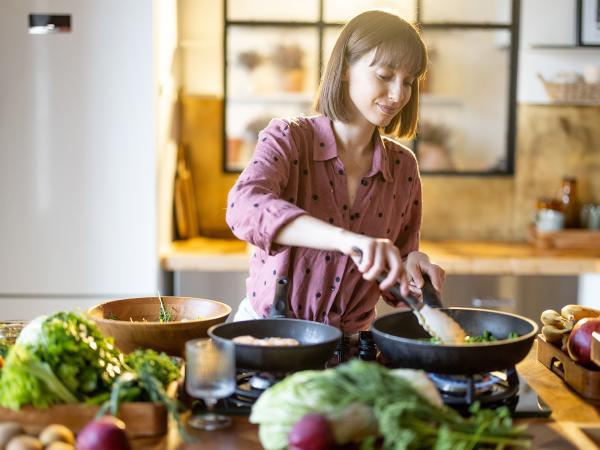 Une personne cuisine devant une plaque de cuisson, mélange des légumes dans une poêle, entourée sur le plan de travail de lég
