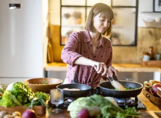 Une personne cuisine devant une plaque de cuisson, mélange des légumes dans une poêle, entourée sur le plan de travail de lég